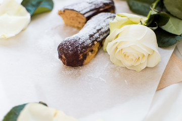 French eclairs with custard sprinkled with powdered sugar and white roses, petals, green leaves on light background