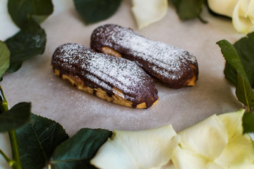 French eclairs with custard sprinkled with powdered sugar and white roses, petals, green leaves on light background