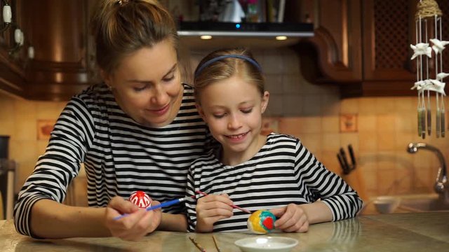 Beautiful caucasian mother and child daughter in striped dresses paint easter eggs having family fun sitting at table at home cosy wooden kitchen