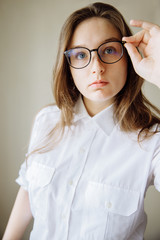 A beautiful brunette girl looks into the frame and adjusts her glasses. The girl is wearing a white shirt with pockets. The girl's hand touches the rim of her glasses. Glasses for the nearsighted.