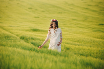 Fototapeta na wymiar pregnant woman in white dress with wreath of flowers on head is relaxing outdoors in grass field. motherhood concept, selective focus