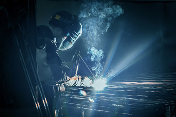 A masked working man is doing welding work on metal structures in a factory or industrial enterprise