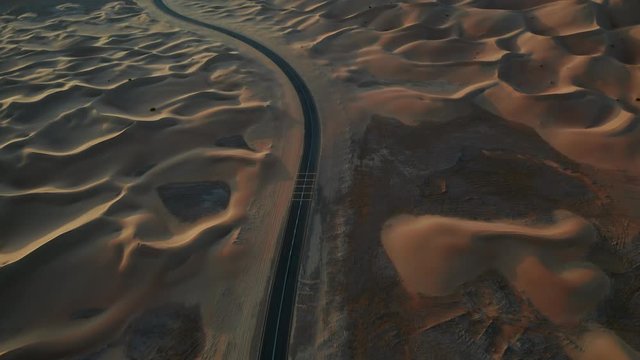 Aerial shot of an empty road in Abu-Dhabi Liwa desert region. Sunrise time with warm vivid colors. Drone flying forward with camera tilts up from road to sand dunes