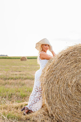Beautiful tanned girl in white lace dress is standing near haystack at sunset in the summer. Happy, smiling woman in straw hat having fun, posing at the field. Concept of countryside lifestyle