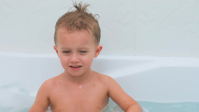 A little boy with sticking hair takes a bath. The child plays in the water, takes the red little ball and throws it into the camera. The boy laughs and looks happily at his mother.