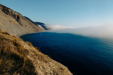 The edge of the juniper forest of Utrish Nature Reserve over the blue sea over which dense fog spreads and finds on high coastal mountains.