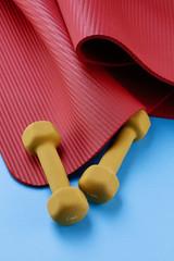 Two yellow dumbbells and a red gymnastic rug/fitness mat on a blue background. Training equipment