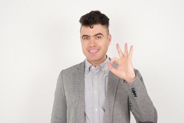 Glad attractive man shows ok sign with hand as expresses approval, has cheerful expression, being optimistic. Standing against gray wall.