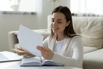 Smiling young woman read pleasant news in paperwork letter at home, happy millennial girl open envelope, get good positive response or message in postal paper correspondence or notice