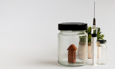 Stay home concept. A house in a transparent jar. Next to the jar is a syringe with medicine and a potted flower in the background. Coronavirus COVID-19 background. Quarantine background. Conceptual. 