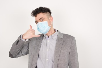 Attractive young male wearing medical mask in casual clothes imitates telephone conversation, keeps hand near ear as if holding mobile phone, has confident facial expression. Call me!