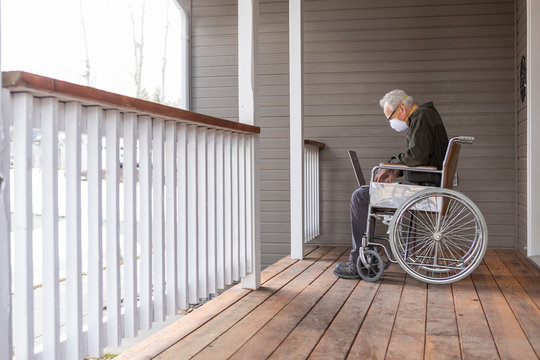 Senior man in wheelchair wearing protective mask to prevent†coronavirus†transmission using laptop on porch