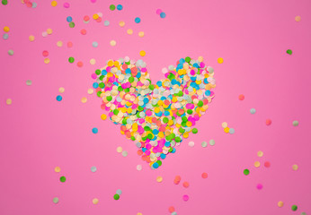 Heart shape made of colorful confetti. Pink backdrop with scattered paper circles. Beautiful decor for the party. Festive backgrond for love story. Valentine's day symbol. View from above. Flat lay