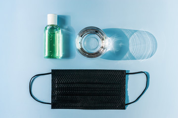 Healthcare Black medical mask, a glass of water and an antiseptic gel on a blue background with hard shadows, top view