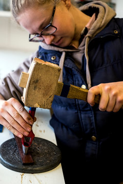 Female saddler standing in workshop, using hand tool and wooden mallet, punching holes into leather strap.