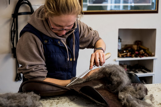 Female saddler standing in workshop, stuffing leather saddle with horse hair.