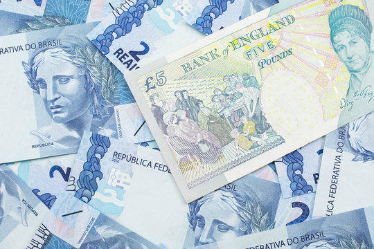 A close up image of a multicolored five pound note from the United Kingdom on a background of Brazilian two reais bank notes in macro