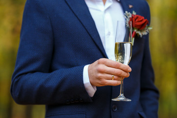 Groom in blue suit with red boutonniere hold in his hands wineglasses with champagne. Wedding bounquet and celebration