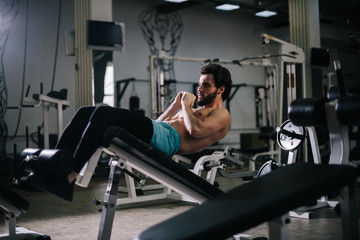 Fototapeta premium Sportive bearded young man with muscular wiry naked torso doing abdominal exercises on floor with black mats during sport workout training in modern dark gym. Concept of healthy lifestyle.