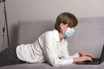 Concentrated young woman wearing medical protective face mask, lying on a grey couch and working on a laptop. COVID-19 pandemic, working at home safely  concept