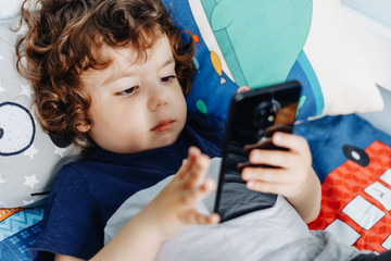 Wow, I like that phone. Baby with smartphone. Boy sitting in bed and playing with mobile phone....