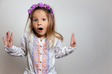 Portrait of pretty little girl in white dress and purple wreath on gray isolated background . Wonder and wow emotion concept