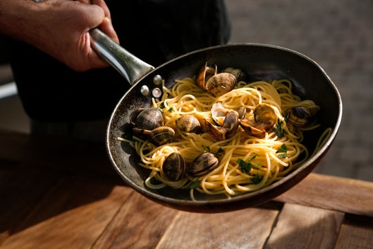 padella con spaghetti alle arselle tenuta da una mano  , sullo sfondo  tavolo di legno