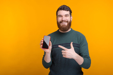 Happy man wearing beard is holding his phone and pointing at it on yellow background