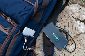 Modern smartphone are charging from power bank lies next to the backpack on a wooden stump. Modern technology concept.