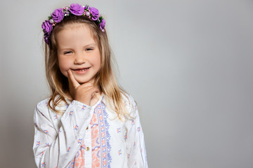 Portrait of pretty little girl in white dress and purple wreath on gray isolated background.  Tricky and sly emotion concept