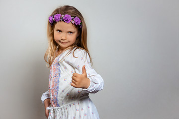 Portrait of pretty little girl  in white dress and purple wreath on gray isolated background. Ok and super emotion concept