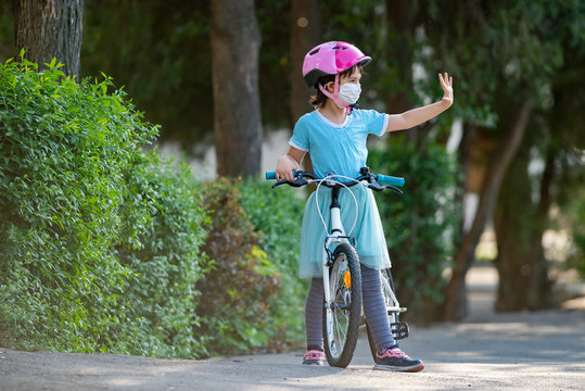 Cute Little Girl With Mask On Face Ridding A Bike And Salute Other Kids From Distance.