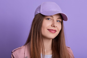 Head shot of attractive cute charming smiling Caucasian female wearing cap, looking directly at camera, posing isolated over lilac studio background, looks happy and confident,has long beautiful hair