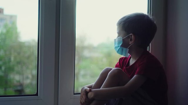 Sad boy in medical mask sits on windowsill and looks out window, self quarantine. View from street. Patient isolated to prevent infection of coronavirus, Covid-19 epidemic, pandemic. Stay home.