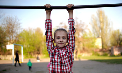 Fototapeta premium Little cute girl in a plaid shirt hangs on the horizontal bar at the playground