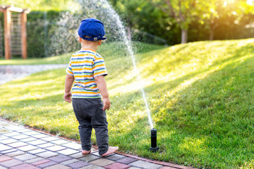 Cute adorable caucasian blond barefeet toddler boy in cap walking at home backyard near sprinkler automatic watering system lawn in garden. Child little helper playing gardening at summer outdoors - obrazy, fototapety, plakaty
