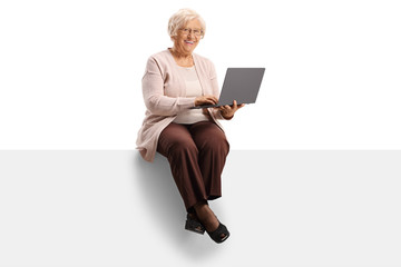 Happy elderly woman sitting on a blank panel and using a laptop computer