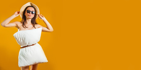 beautiful brunette woman in a white pillow dress and summer hat spreads against a yellow studio background. Fashionable girl. Pillow call due to stay at home isolation. Coronavirus quarantined.