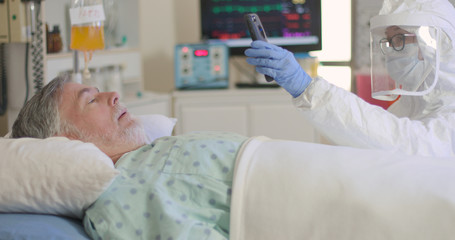 A nurse or doctor in full PPE helps a hospitalized patient with COVID19 keep in touch with his family via a video conference on a cell phone.