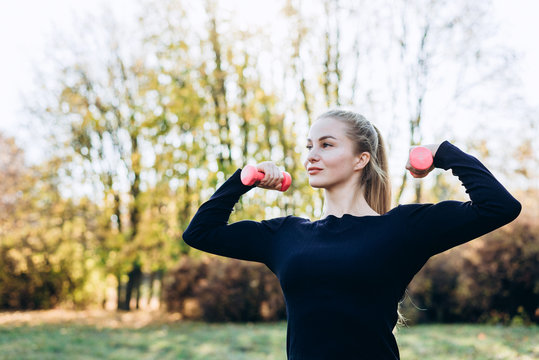 Beautiful young girl posing with dumbbell, outdoors, looking away.