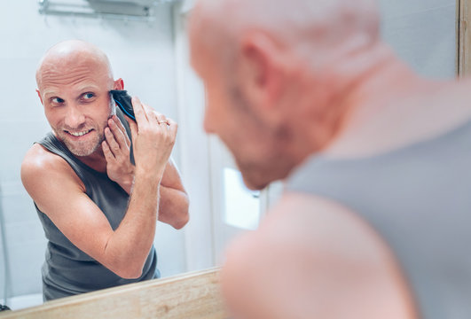 Hairless man fixing his beard looking in bathroom mirror using an electric rechargeable Beard Trimmer. Everyday body and skincare routine treatments concept image.