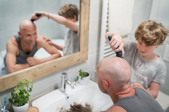 Teenager son helping his father to trim a bald head gently using electric rechargeable Trimmer in bathroom. Funny home scene and family relatives concept image.