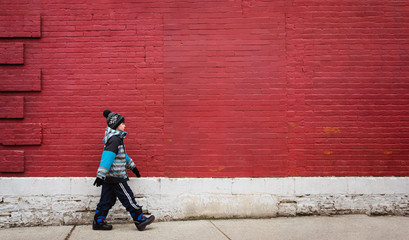 Young boy in winter clothing walking on sidewalk in front of red wall.