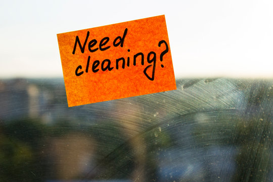 A dirty window in the mud stains, on which a sticker is pasted, with the inscription: need cleaning, and a question mark. A bright orange note. Background behind the glass in the blur.