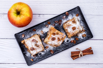 Slices of homemade Apple pie strudel in a plate with ingredients on a rustic white wooden table. Pie with apples, raisins and cinnamon, sweet pastries - Top View