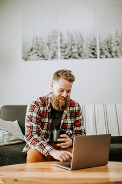 man with beard holds cup of coffee while working on laptop computer