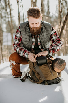 man with beard wearing flannel kneels in snow and adjusts hiking pack