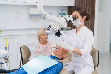 A dentist examines the oral cavity of a woman with the help of a special medical appliance, while her little daughter is watching them