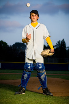 Portrait of a boy standing in white baseball uniform and catchers shin guards tossing a baseball in the air
