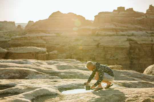 blonde man in camouflage jacket collects water from a puddle in desert